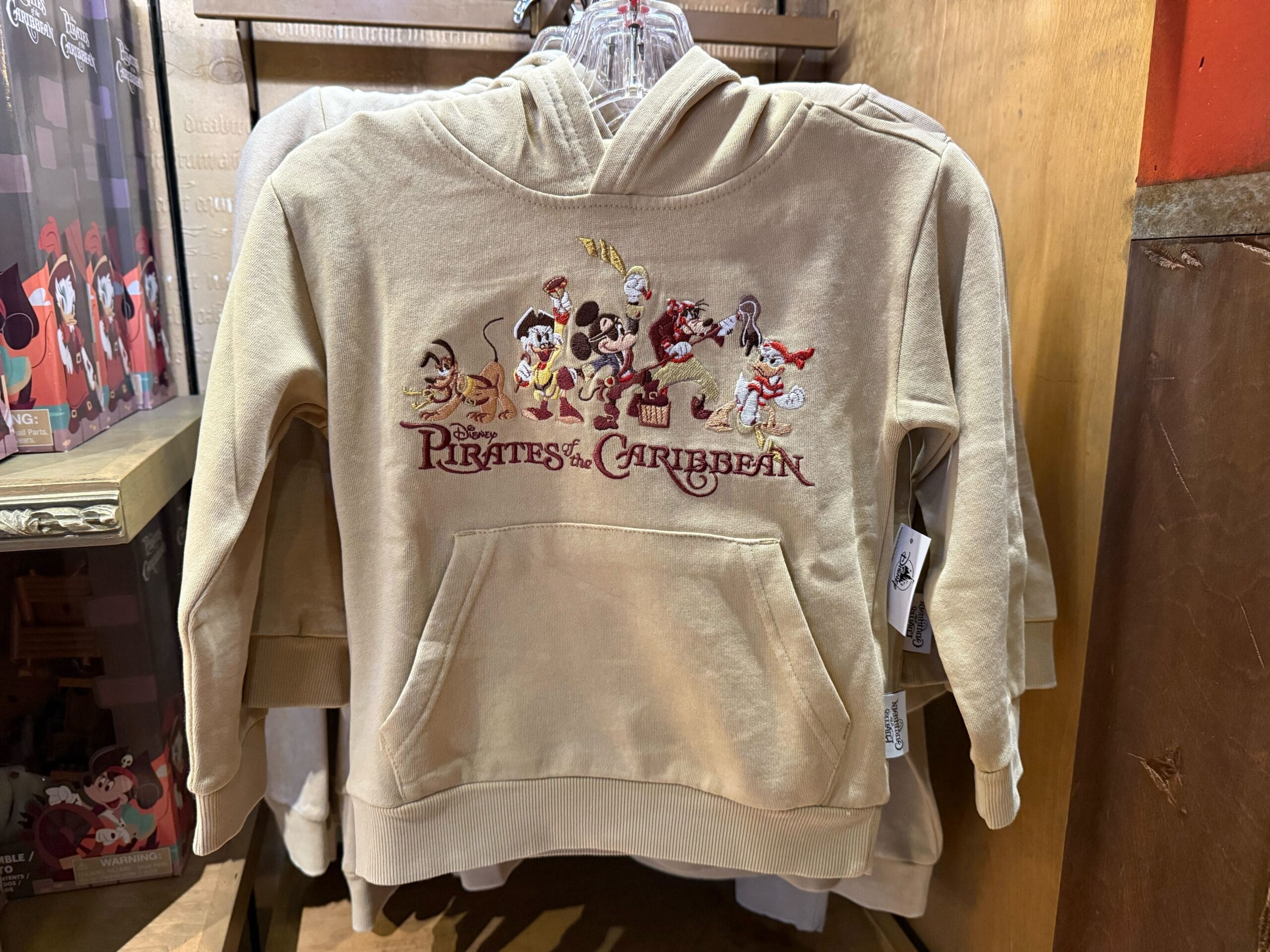 Beige hoodie with cartoon Pirates of the Caribbean characters and logo, hanging on a clothing rack in a Disney park store.