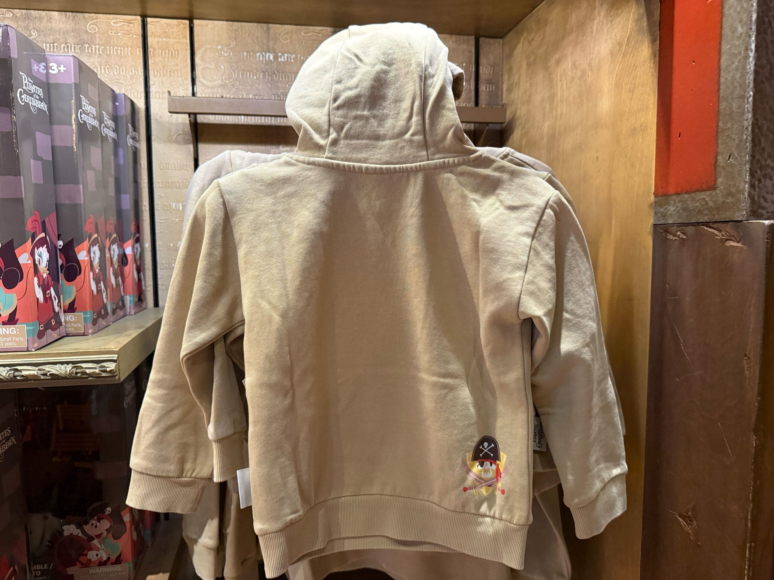 Beige kids' hoodie featuring a small pirate design on the lower back, hanging on a shop rack at a Disney park.