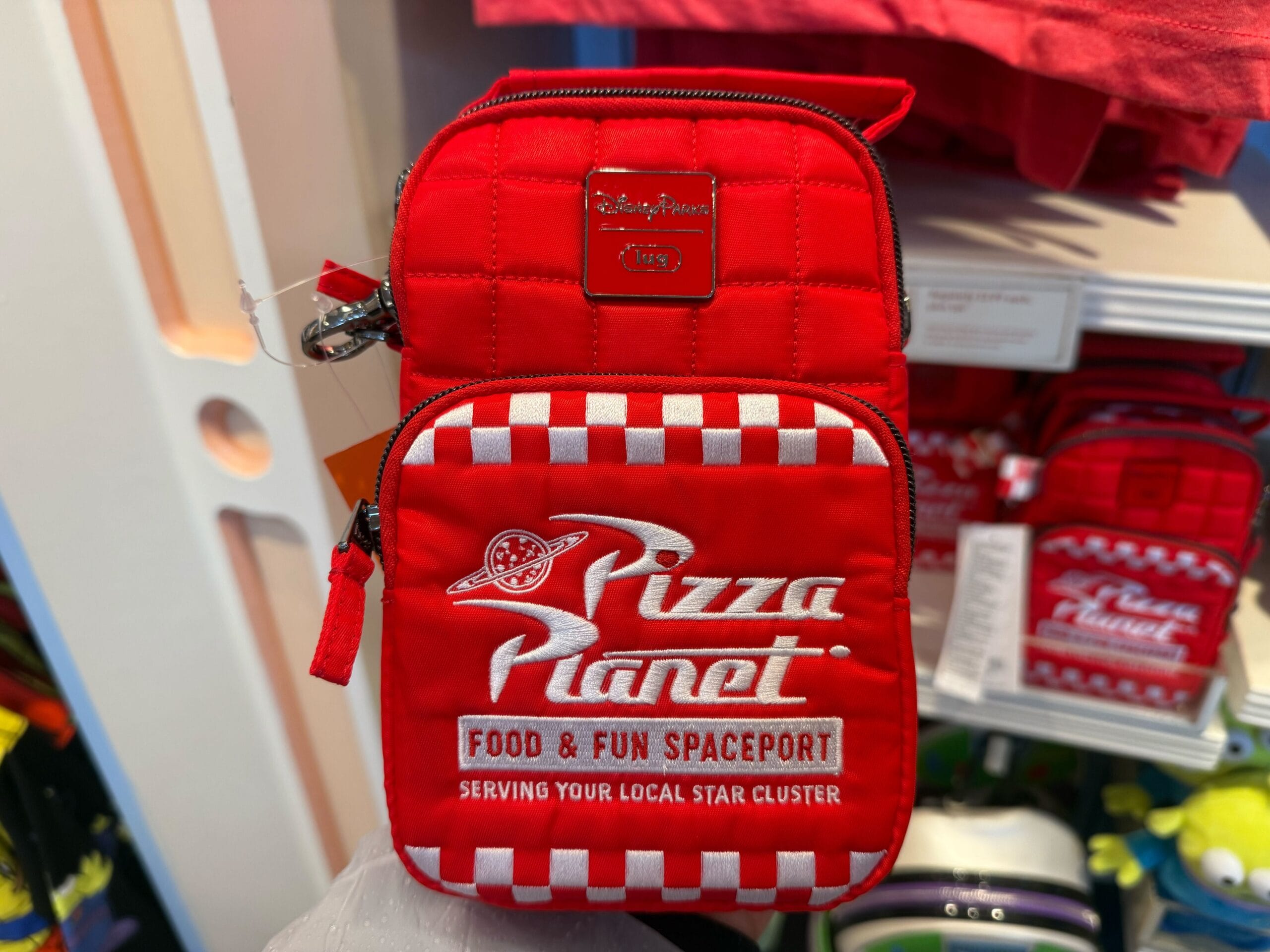 A red "Pizza Planet" bag with white lettering and checkered design, seen in a Disney park shop, reminiscent of Pixar theming.