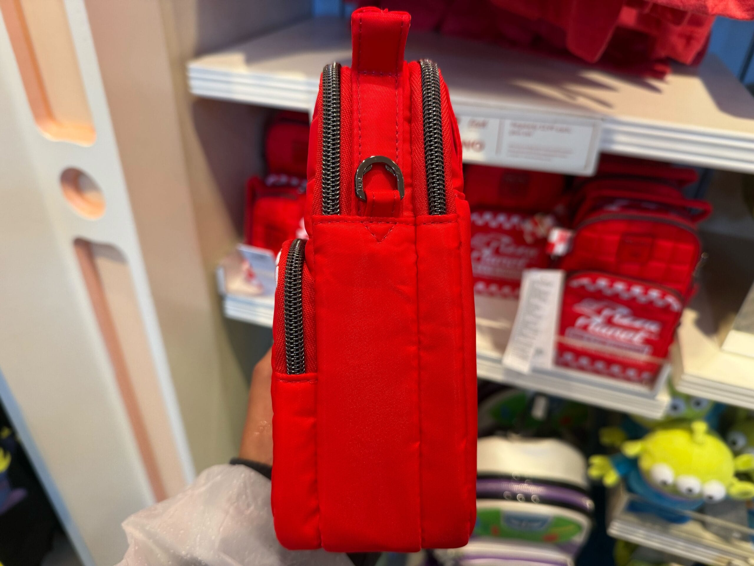 Side view of a small red backpack with silver zippers, seen in a Disney park shop with other vibrant bags behind it.