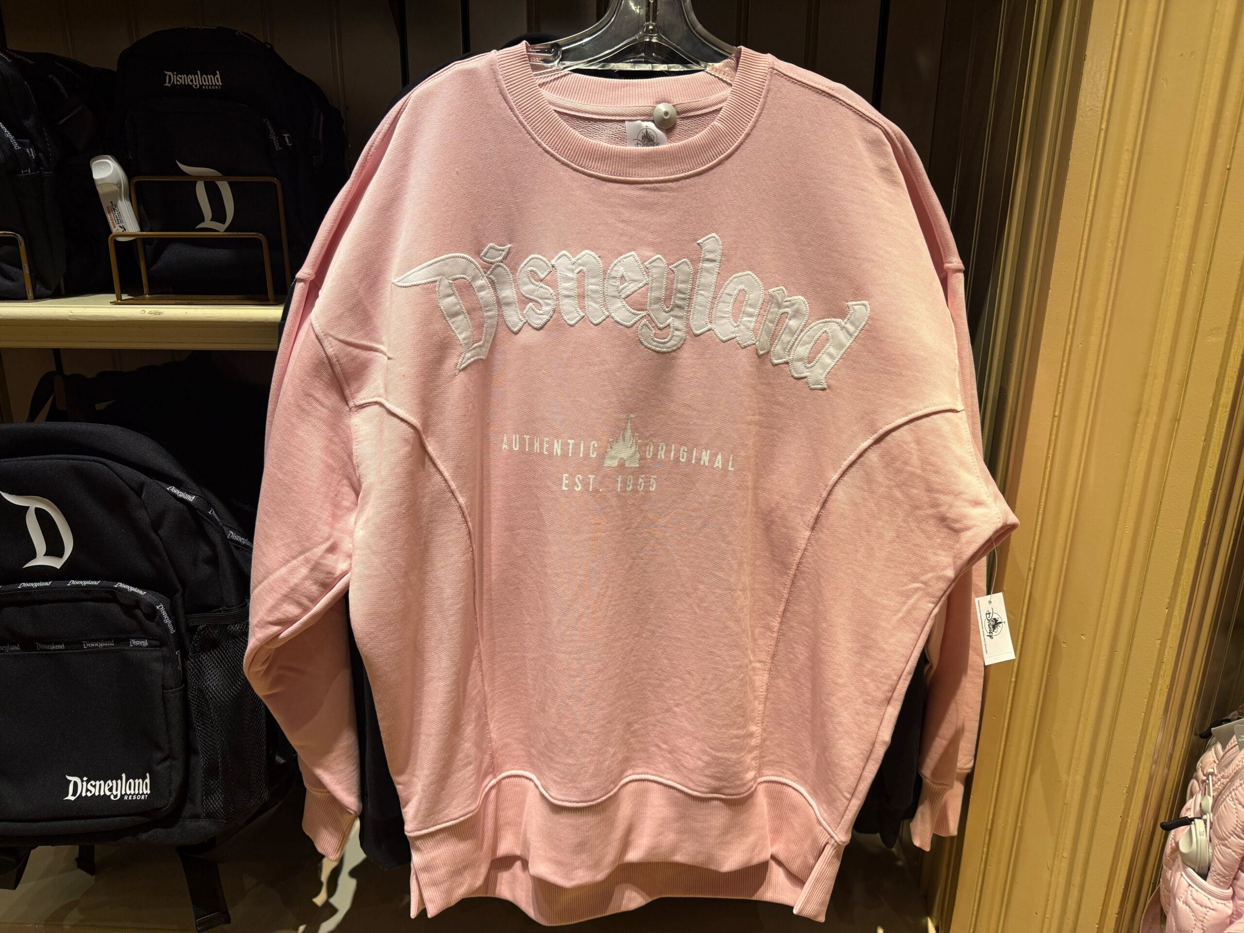 A pink Disneyland logo sweatshirt with white embroidered letters hangs on a rack in a store.