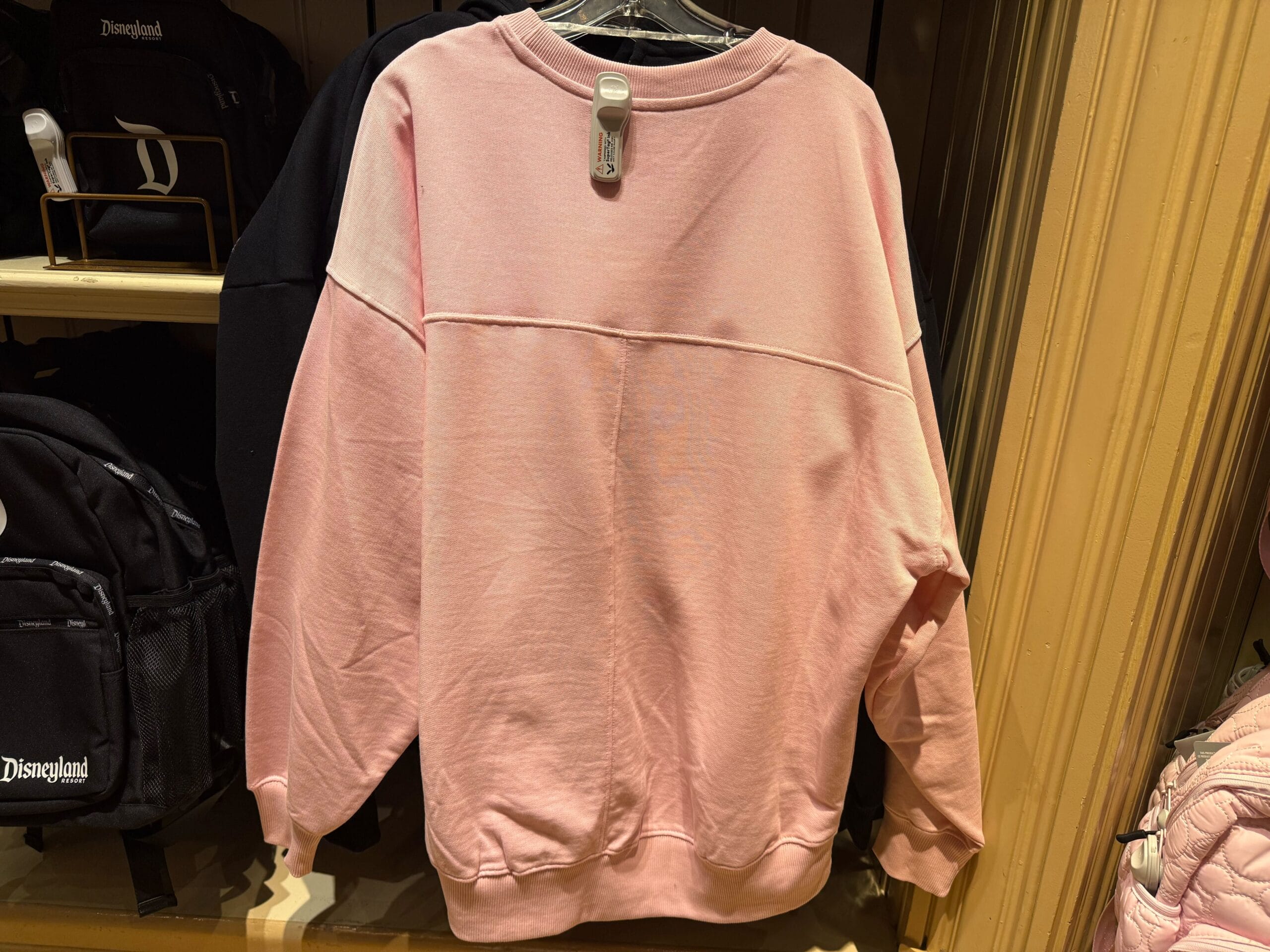 A pink long-sleeve Logo Sweatshirt hangs on a rack in a store, with backpacks displayed nearby.