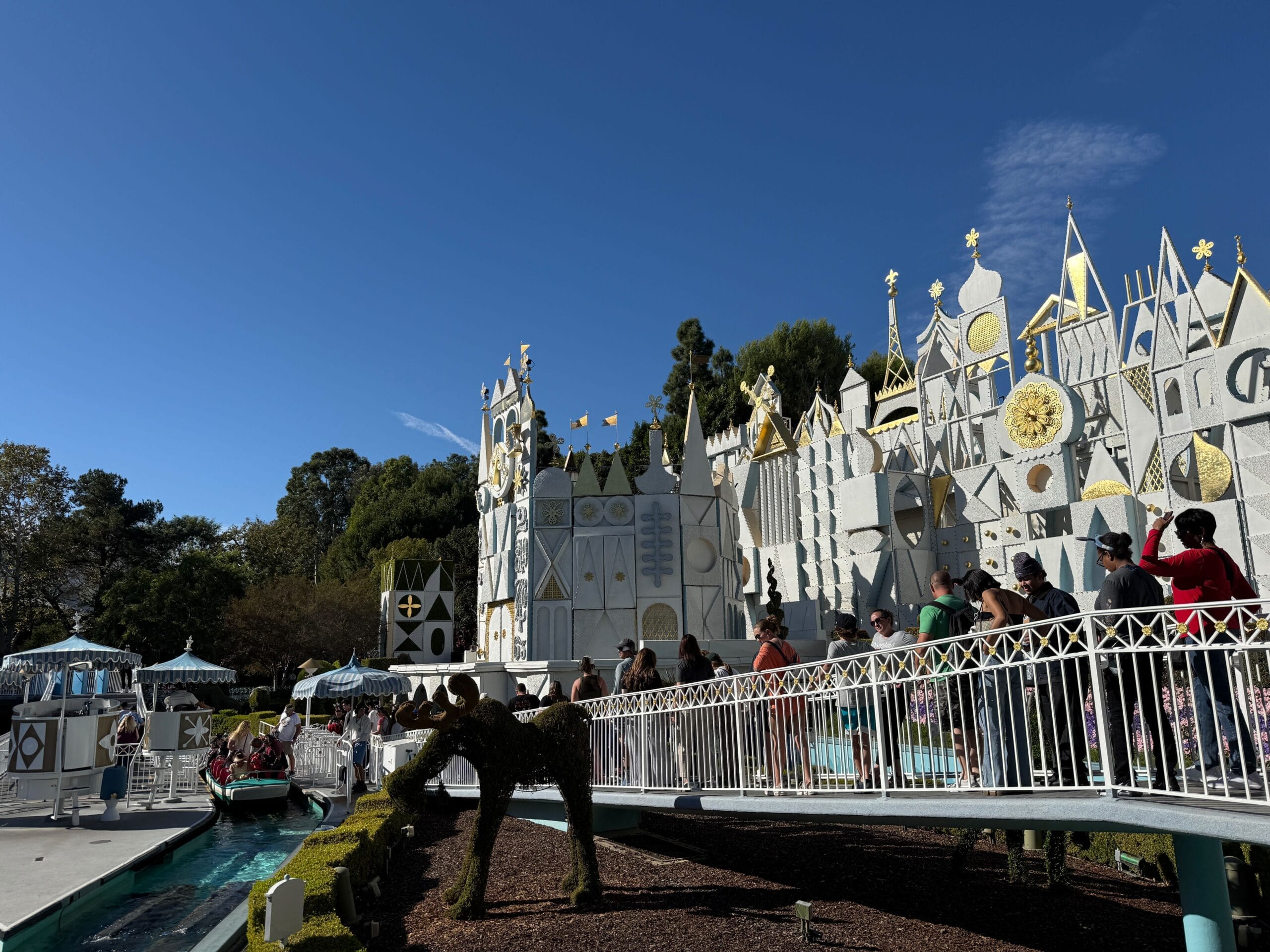 Guests queue outside Disneyland's "It's a Small World" under clear blue skies—no castle in view to compare among Disney Parks.