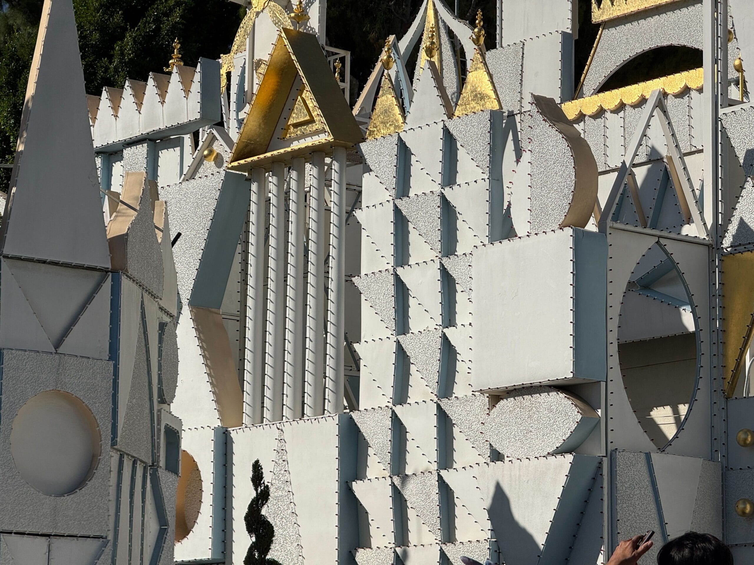 Geometric white and gold facade with whimsical shapes, recalling Disneyland’s it's a small world, crowds below in partial shade.