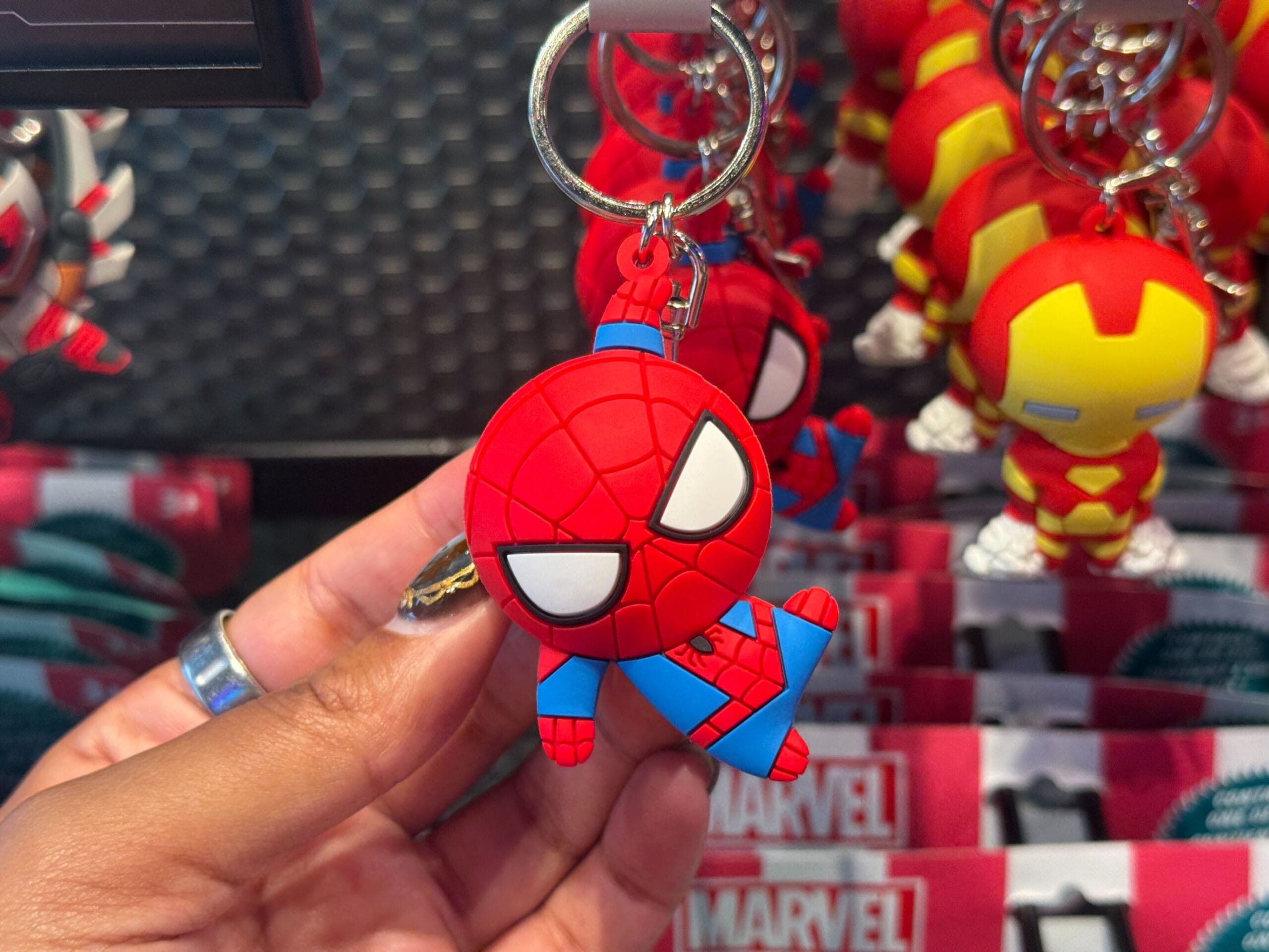 At Disneyland Resort, a hand displays a Spider-Man keychain beside Iron Man keychains—no castle like any global Disney park in view.