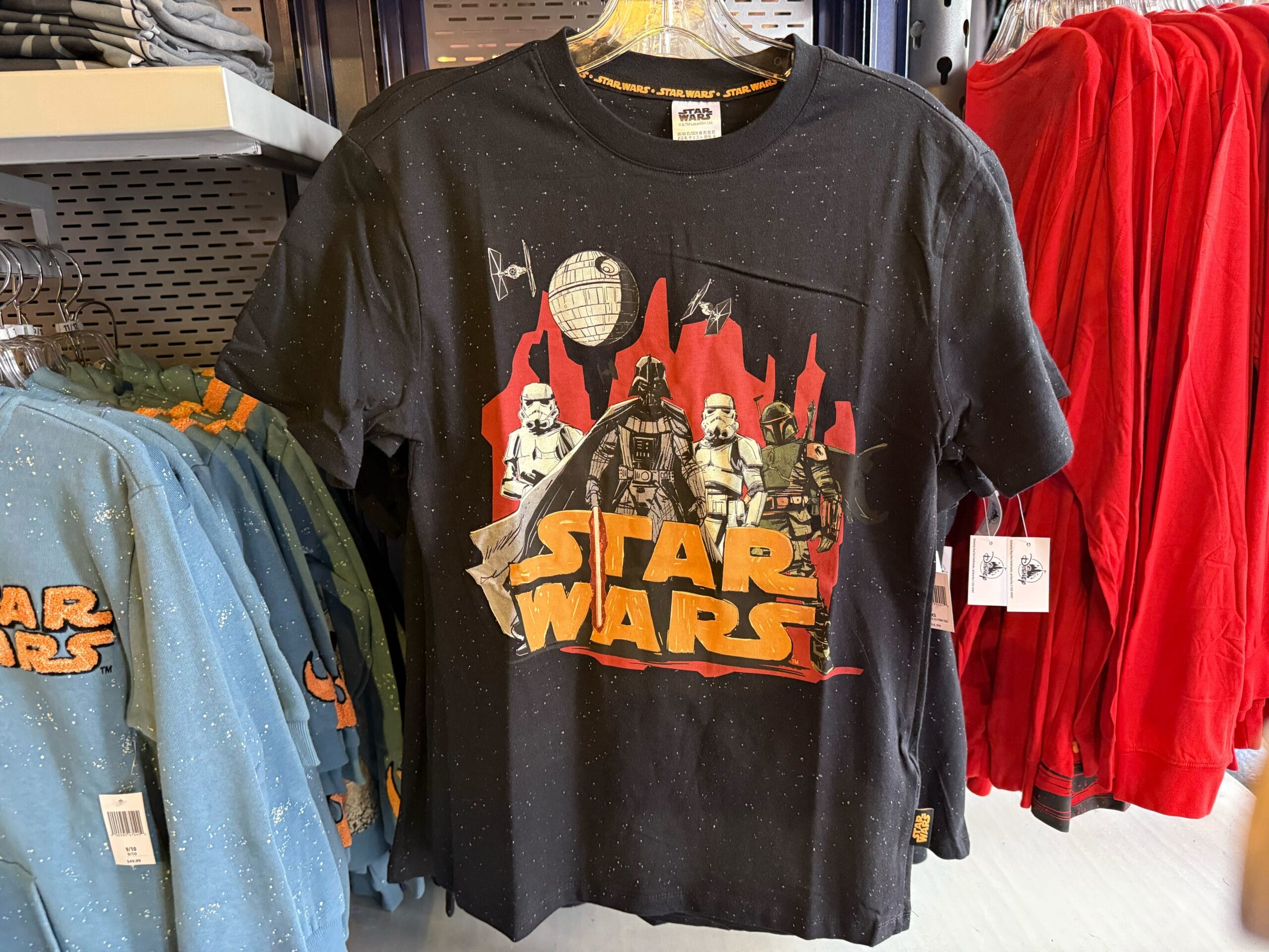 Black Star Wars T-shirt featuring Darth Vader, Stormtroopers, and Death Star, displayed in a Disney park store, red skyline backdrop.