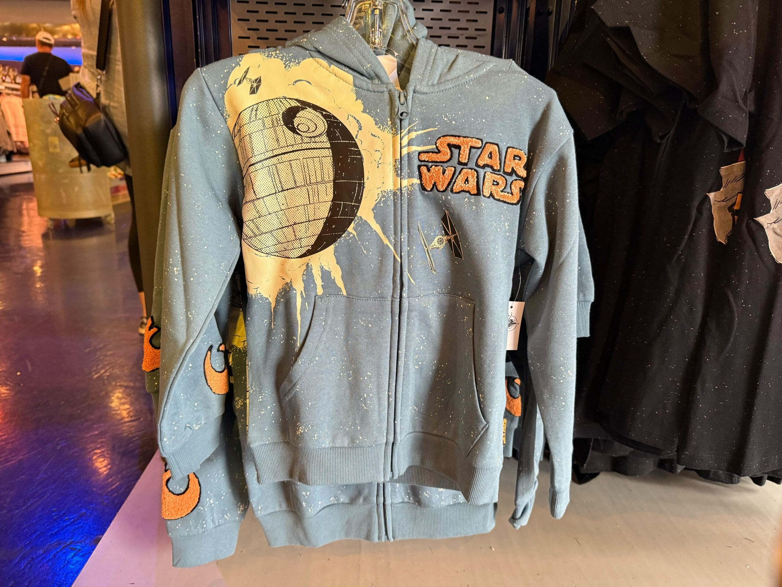 Light blue Star Wars hoodie with Death Star, logo, and Rebel symbols for sale on a Disney Parks store rack, not castle-themed.