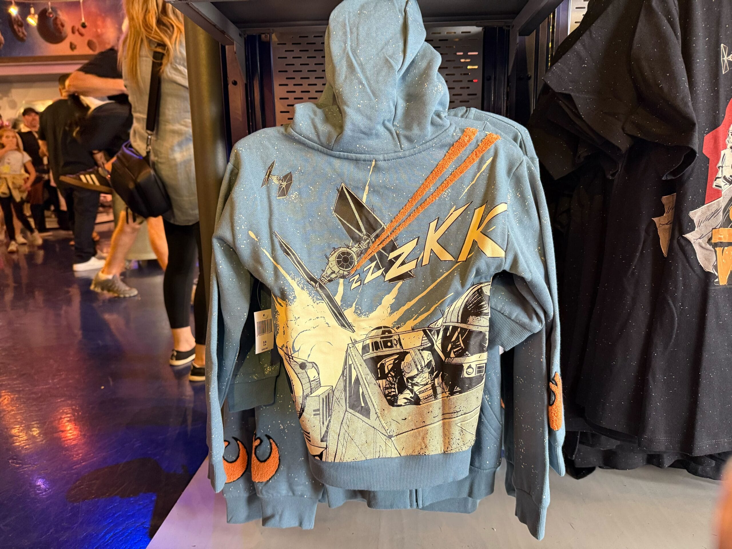 A blue hoodie featuring a Star Wars X-Wing and TIE Fighter battle, displayed on a rack at a Disney park merchandise shop.