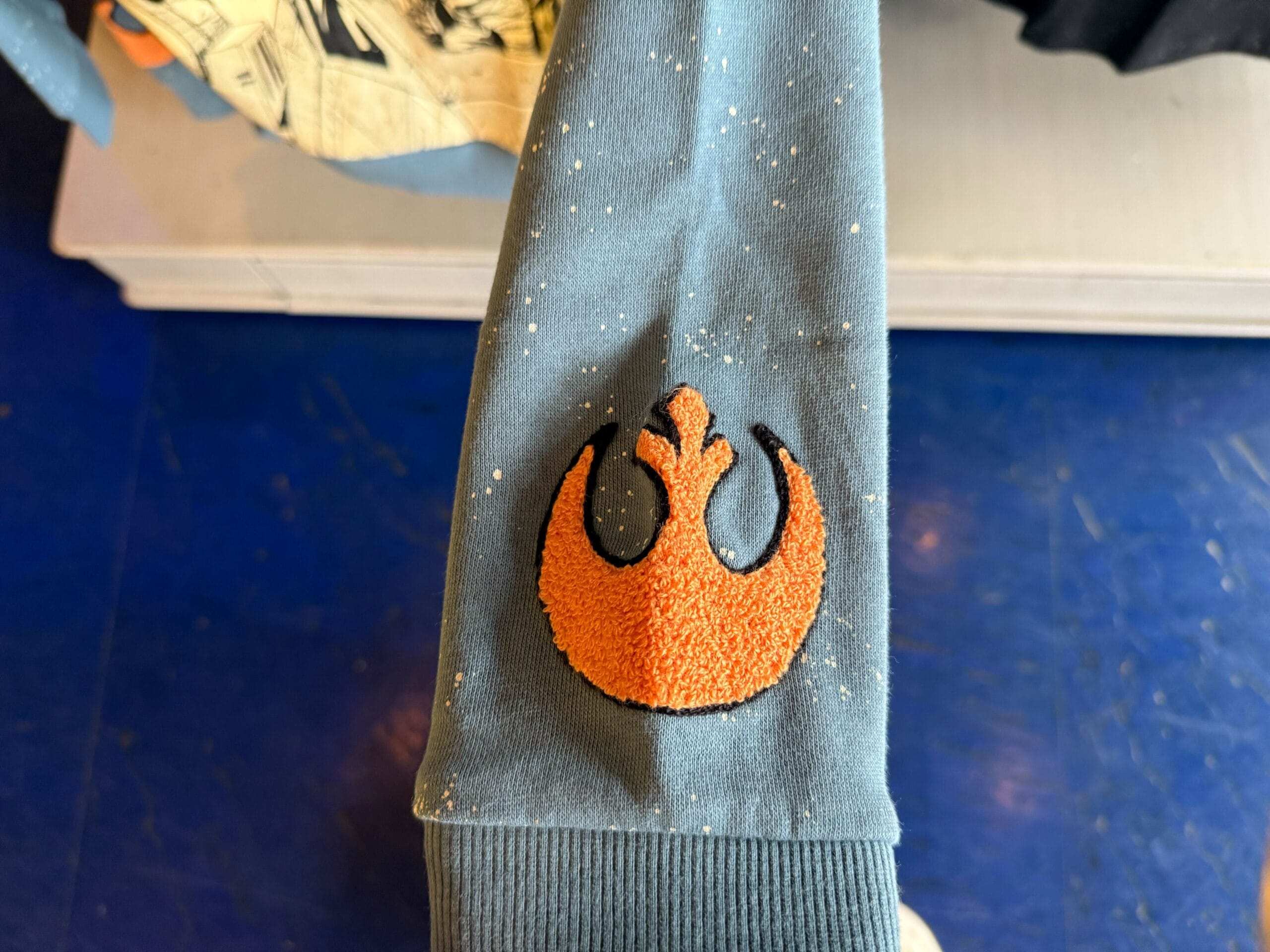 An orange Star Wars Rebel Alliance patch is stitched on a blue, speckled sleeve, perfect for fans at a Disney park.