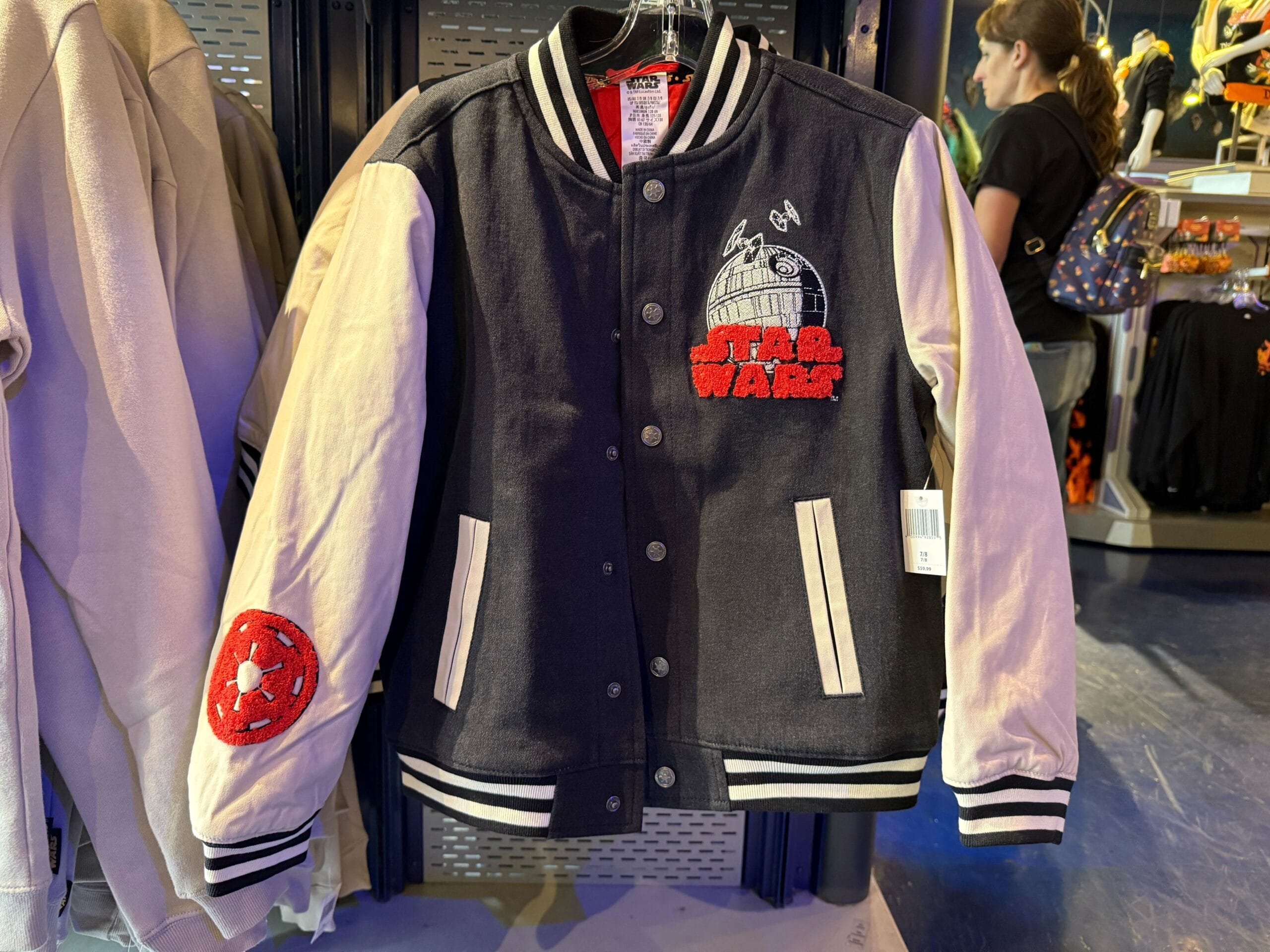 A black and white Star Wars varsity jacket with patches and embroidery hangs on a rack at a Disney park merchandise store.