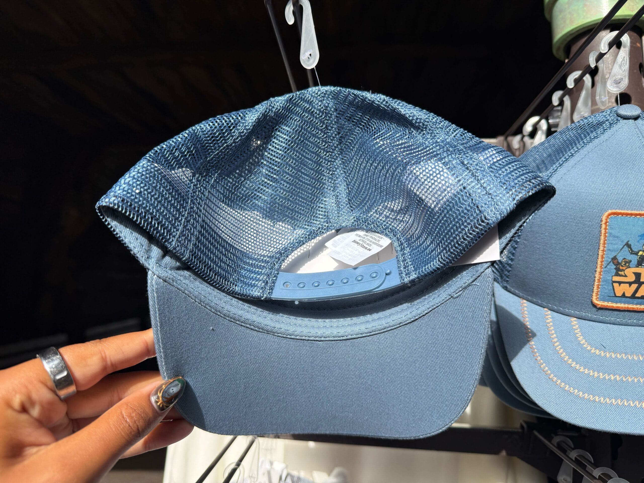 A hand holds a blue mesh-back cap with an adjustable strap and Star Wars tag, offered in bright sunlight at a Disney park.