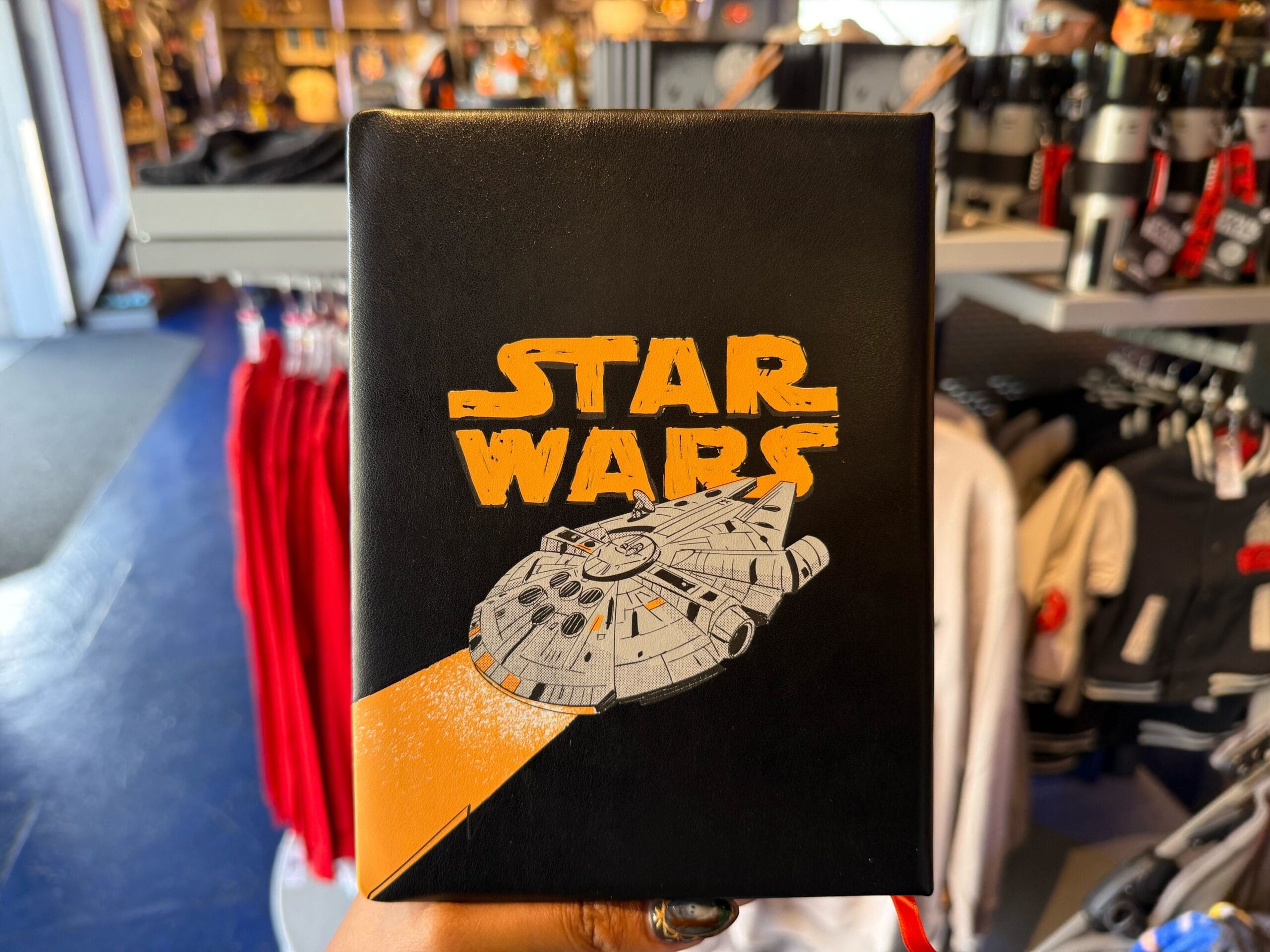 A hand holds a Star Wars Millennium Falcon apparel box in a Disney park store, shelves of clothes behind—no castle in view.