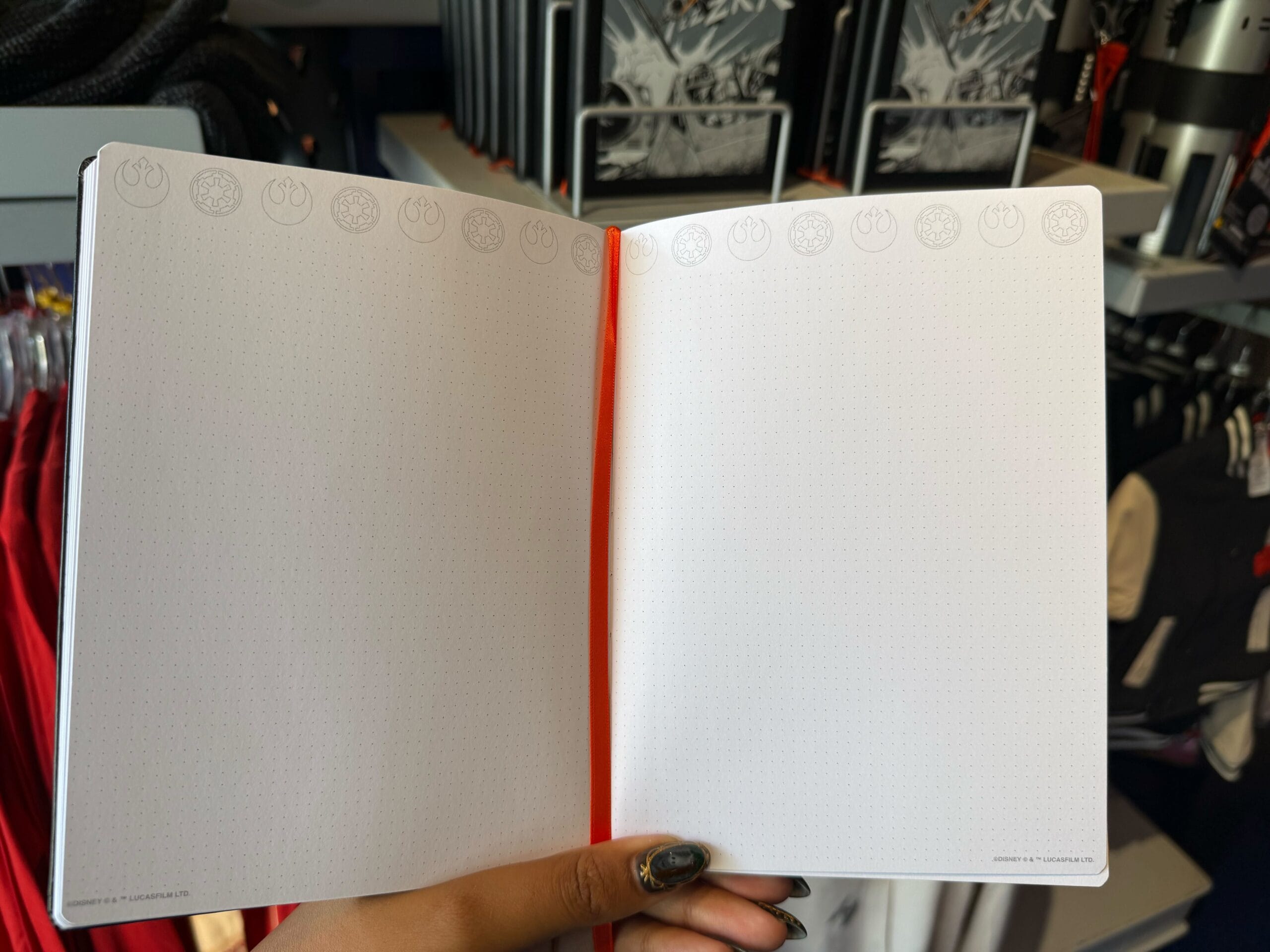 A hand opens a blank Star Wars notebook with Darth Vader; Disney Parks apparel fills shelves behind, no castle in view.