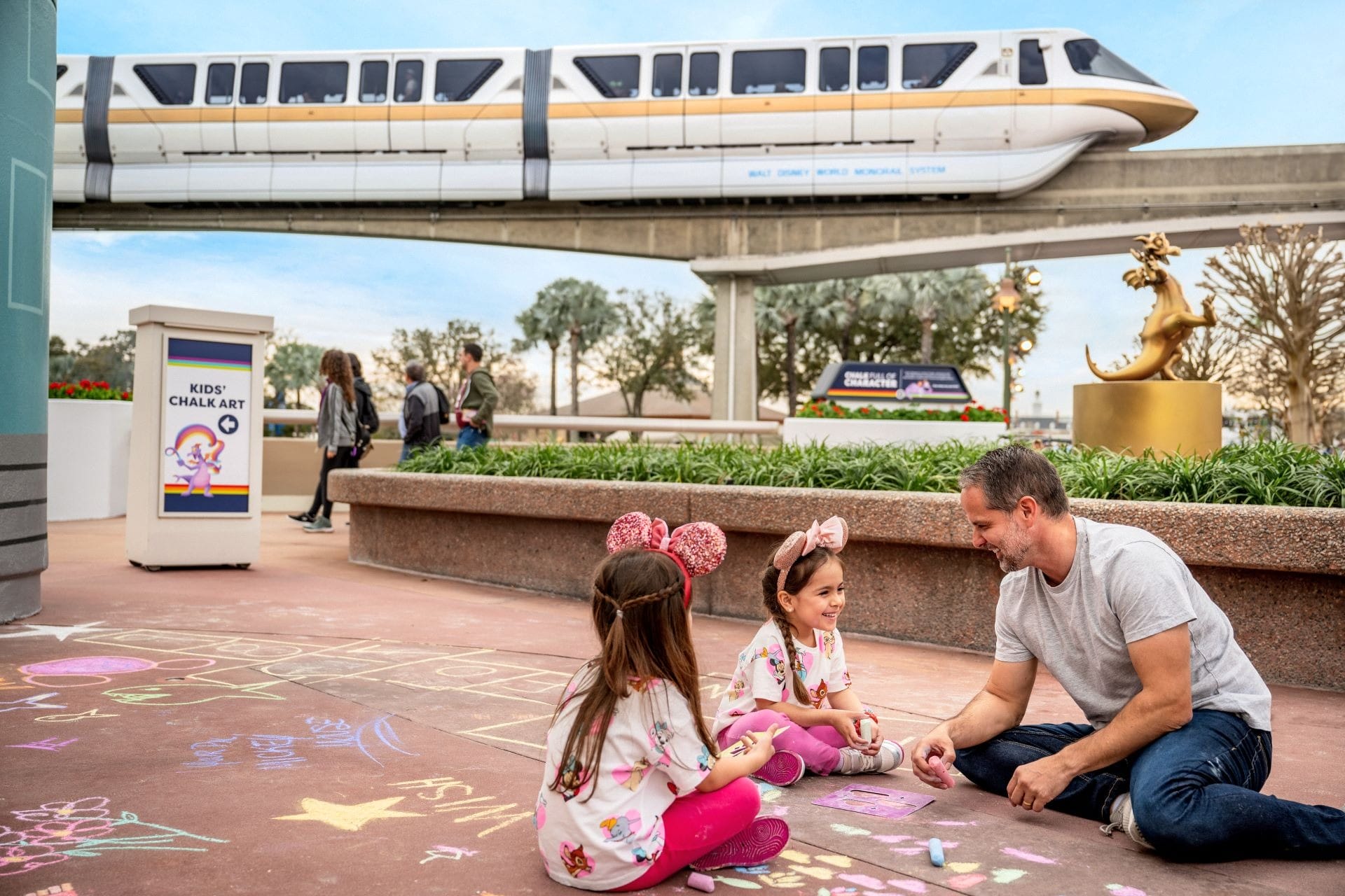 A man and two girls create chalk art at EPCOT’s Festival of the Arts as a monorail glides by, 2026—no castle in sight here.