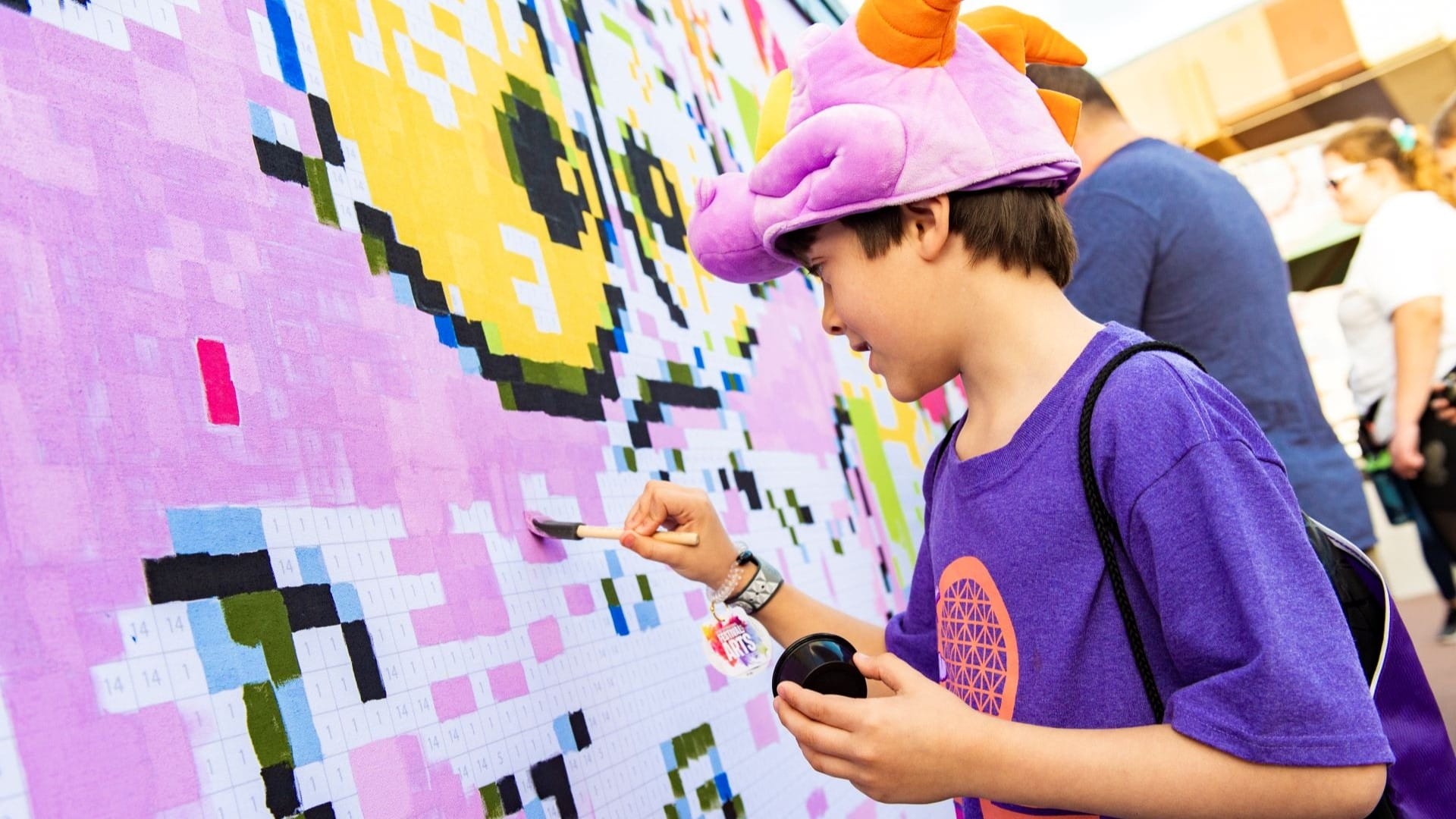 A boy wearing a purple dragon hat paints a color-by-numbers mural at the 2026 EPCOT International Festival of the Arts.