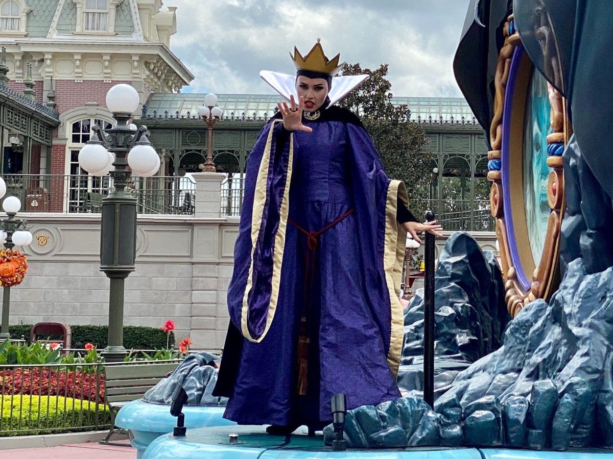 The Evil Queen from Disney Villains poses on a parade float, wearing a purple gown and crown, at a Disney park event.