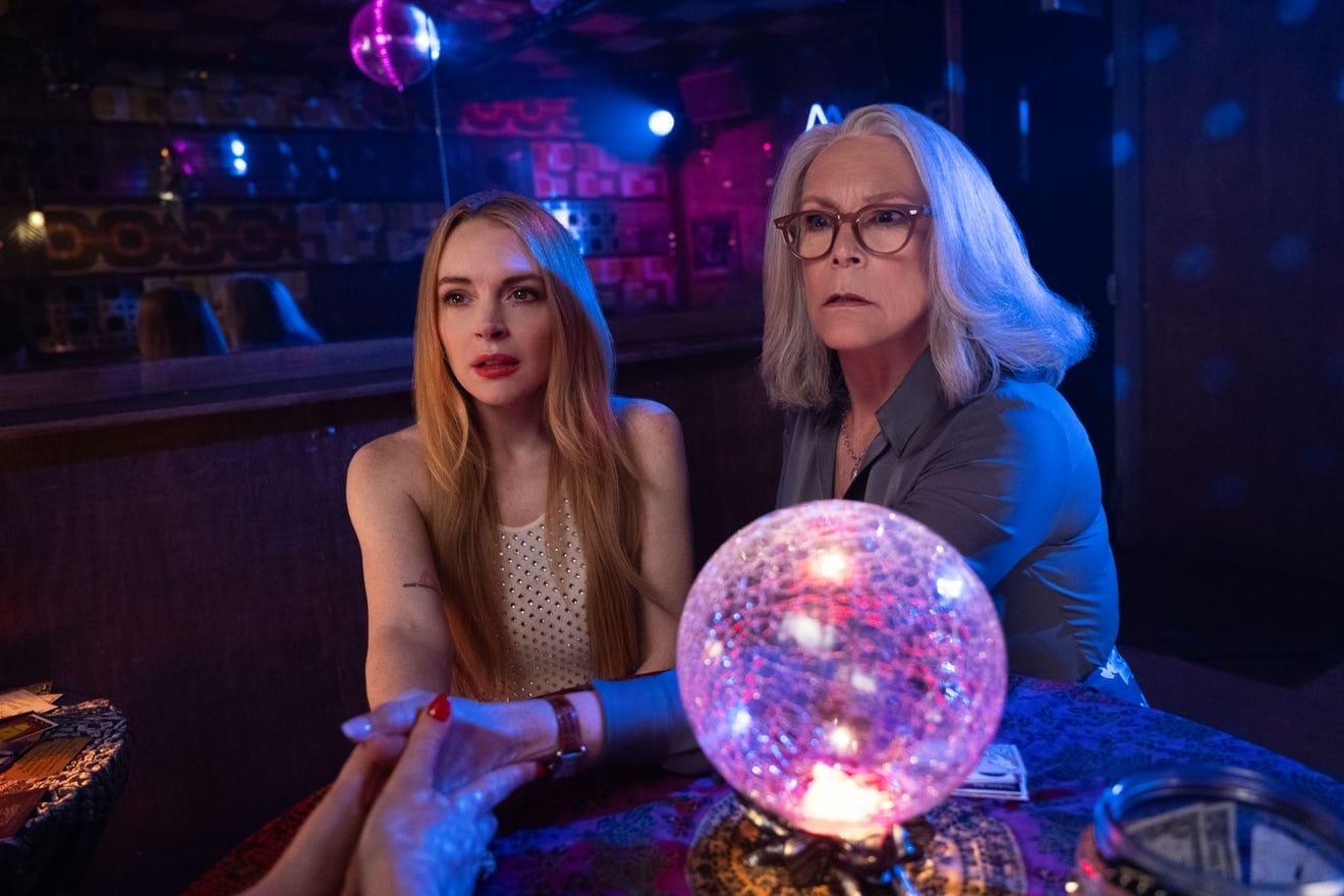 Two women at a table with a crystal ball, holding hands with someone off-screen in a dim, Haunted Mansion-like setting.