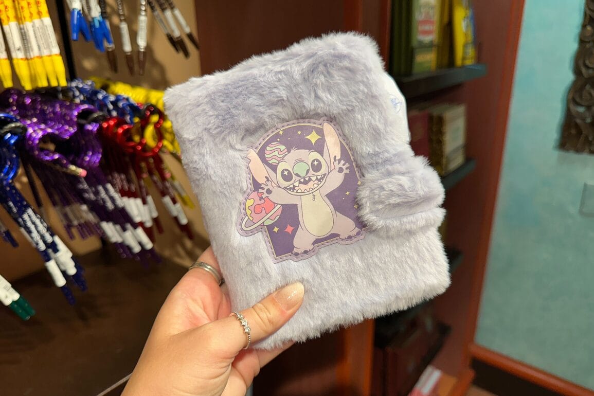 دستی که یک دفترچه یادداشت خاکستری پشمالو با طرح یک شخصیت تعطیلات دیزنی به همراه پالت نقاشی را نگه داشته است، یادآور سبک والت دیزنی ورلد.