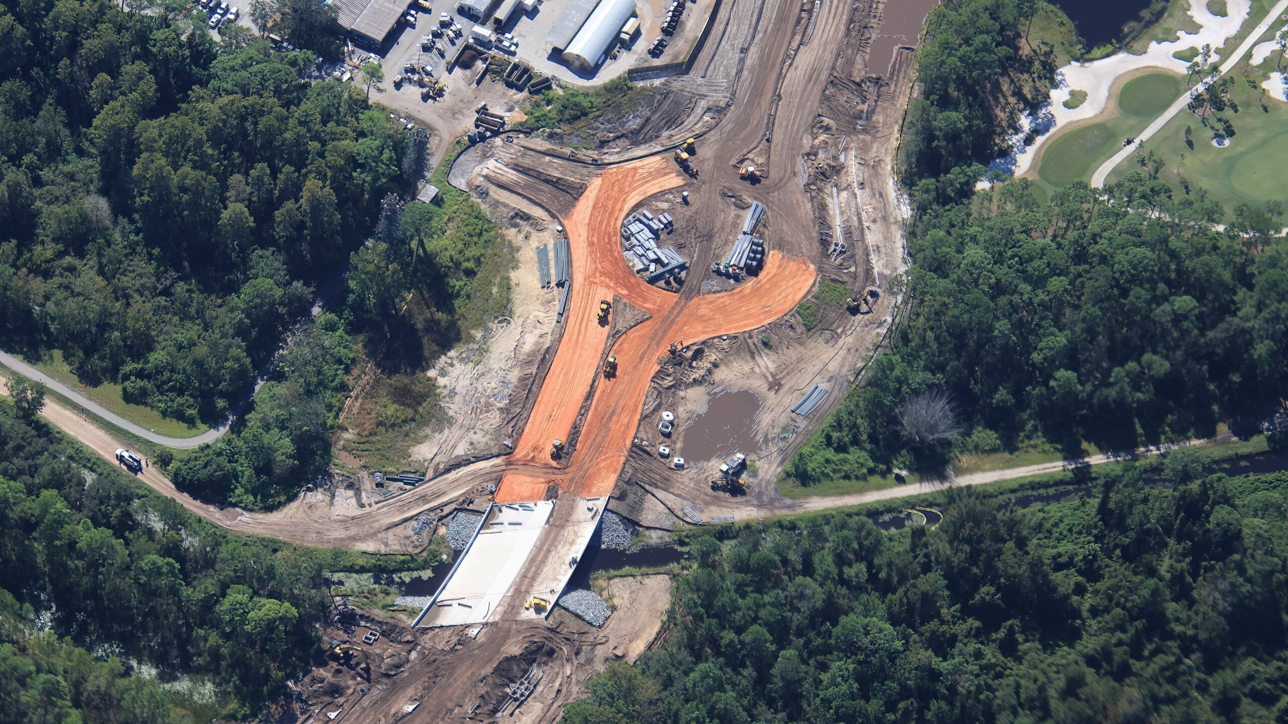 Aerial view of a road construction site with heavy machinery, a bridge, and a visible traffic circle among trees, not near any Disney castle.