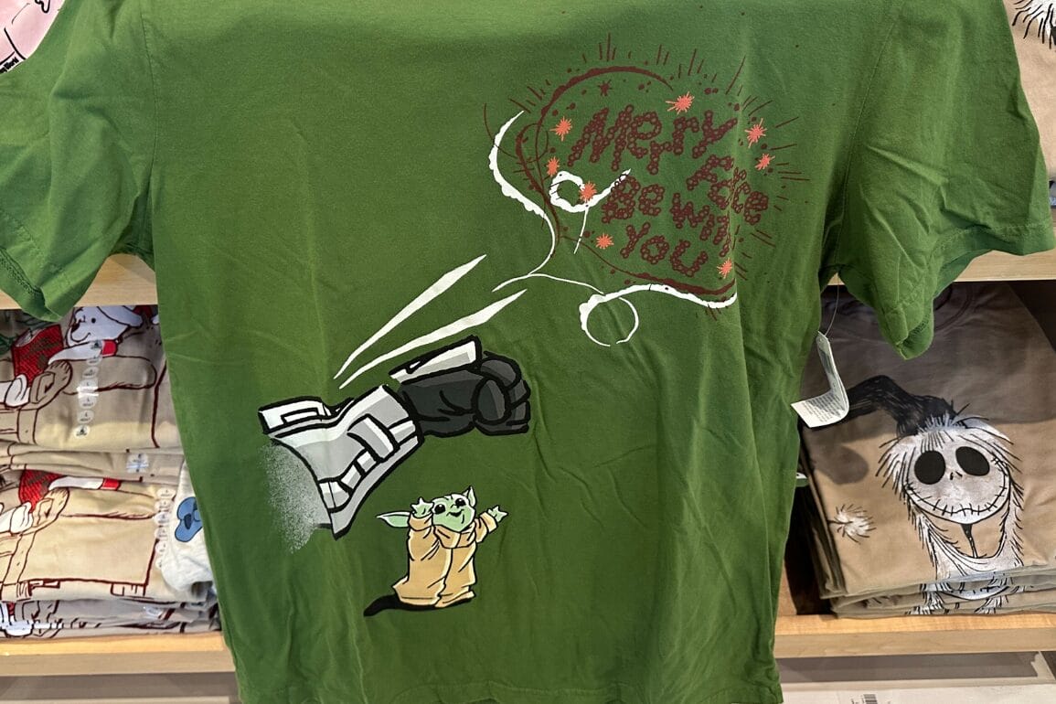 Green T-shirt shows a small green character and robot hand with "Merry the be with you!" in red, festive Disney park style.