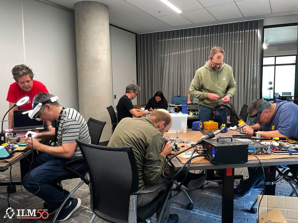 People work on electronics at tables in an office, with a Star Wars ILM 50 logo—no Disney park or castle imagery shown here.