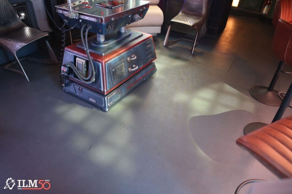 A boxy robot stands on a dark floor in a Lucasfilm employee lounge; ILM 50 logo in the corner, no Disney castle visible.