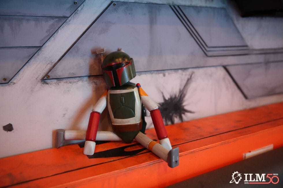 A Boba Fett doll rests by a sci-fi wall in the Star Wars Employee Lounge, ILM 50th logo, evoking Star Wars Galaxy’s Edge vibes.
