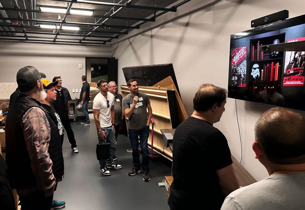 Employees in a lounge watch a man present Star Wars visuals on a big screen, reminiscent of Disney Parks' themed talks.
