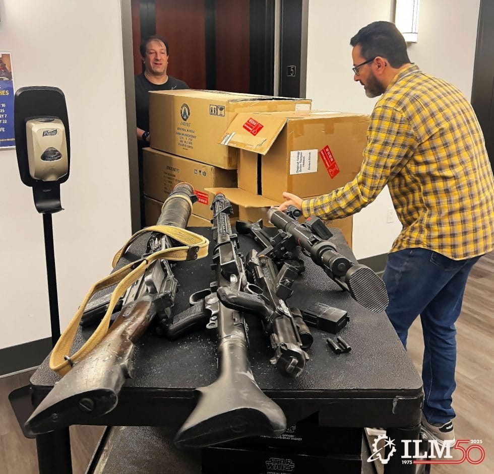 Two men move large boxes while Star Wars prop blasters rest on a black table in an employee lounge, not far from Disney Park props.