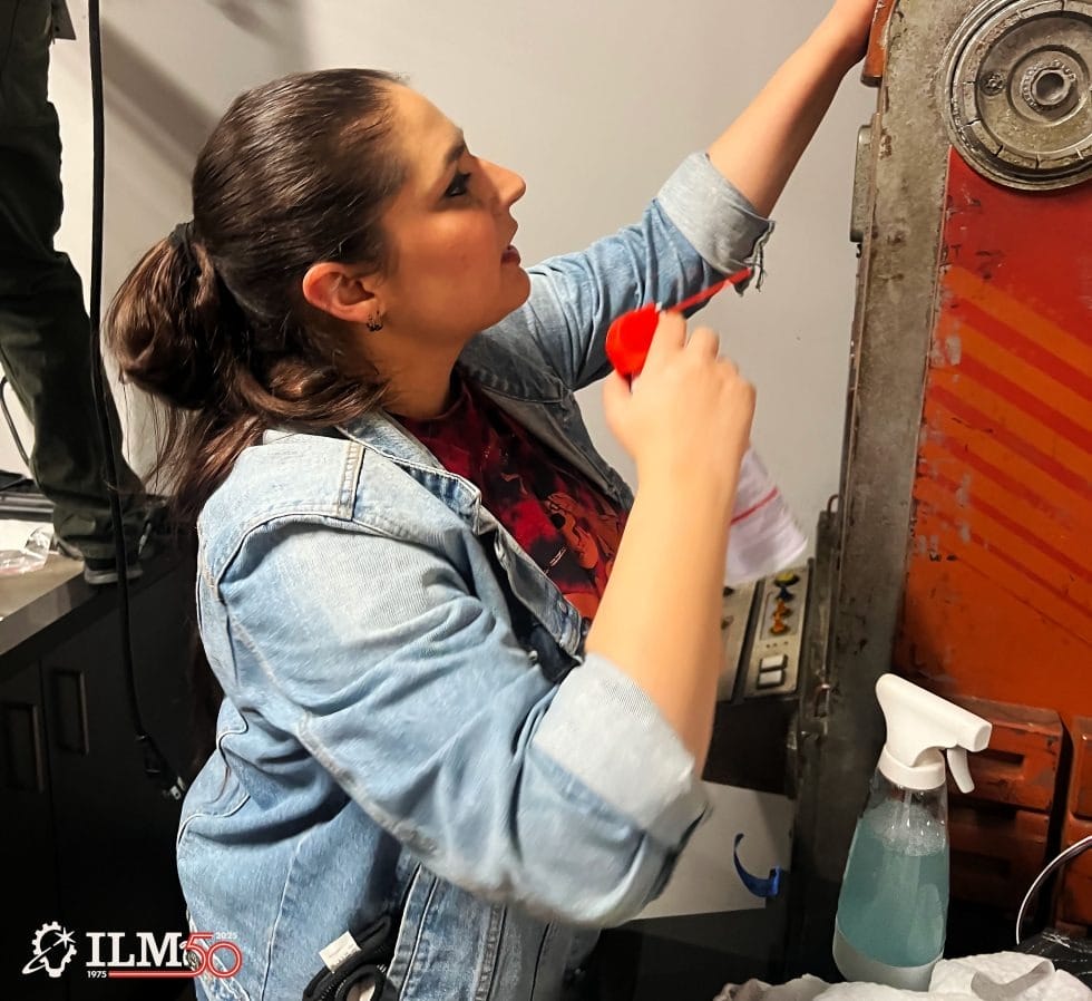 A woman in a denim jacket cleans a large machine with a spray bottle, like backstage staff at Disneyland’s Star Wars attractions.