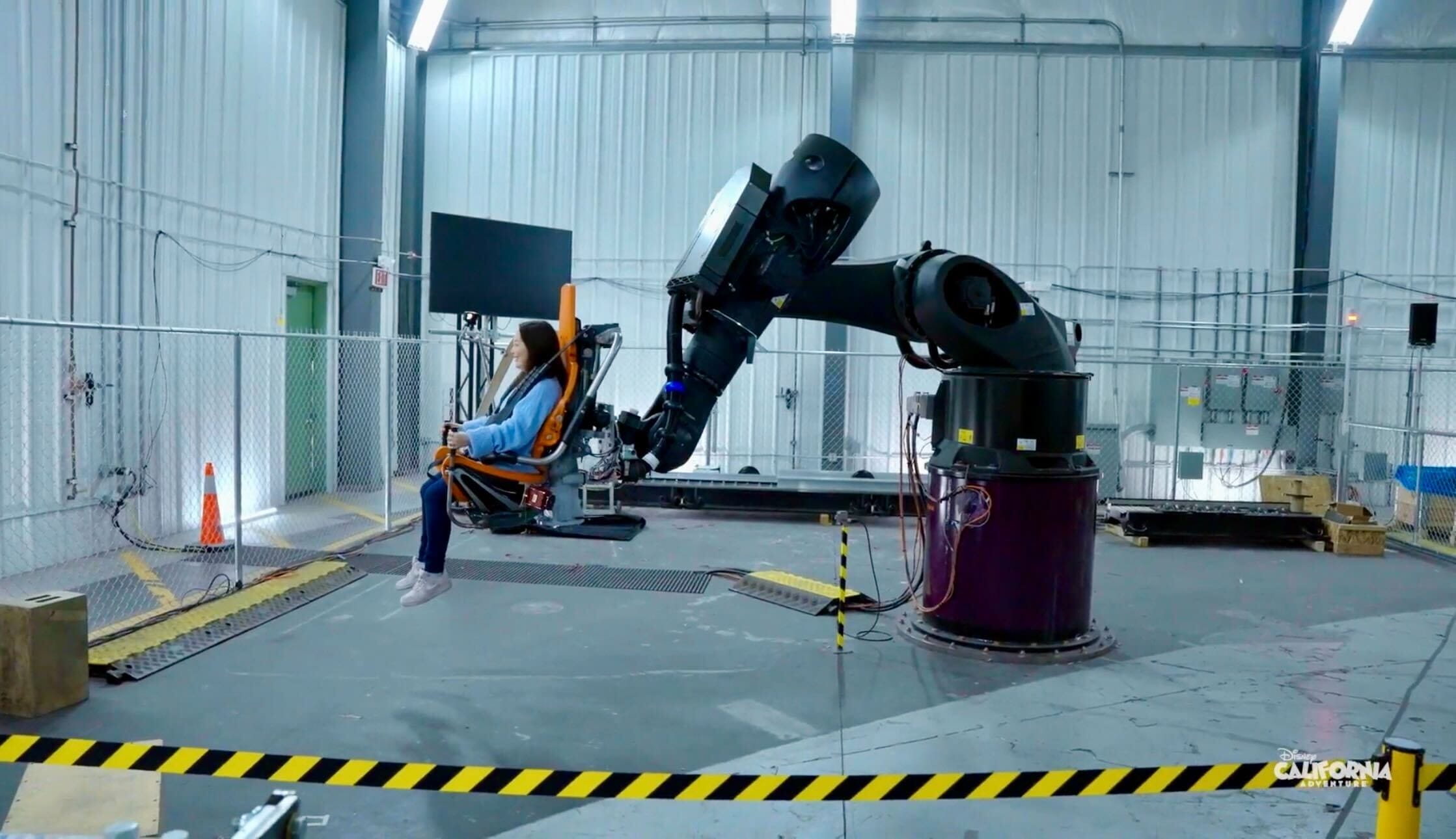 A person sits in a chair on a robotic arm inside the Avengers ride building at Disney, surrounded by safety barriers—no castle visible.