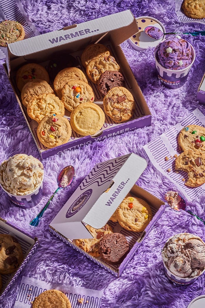 Insomnia Cookies boxes and ice cream tubs on a fluffy purple surface, reminiscent of Disney park treats, with spoons and cups.