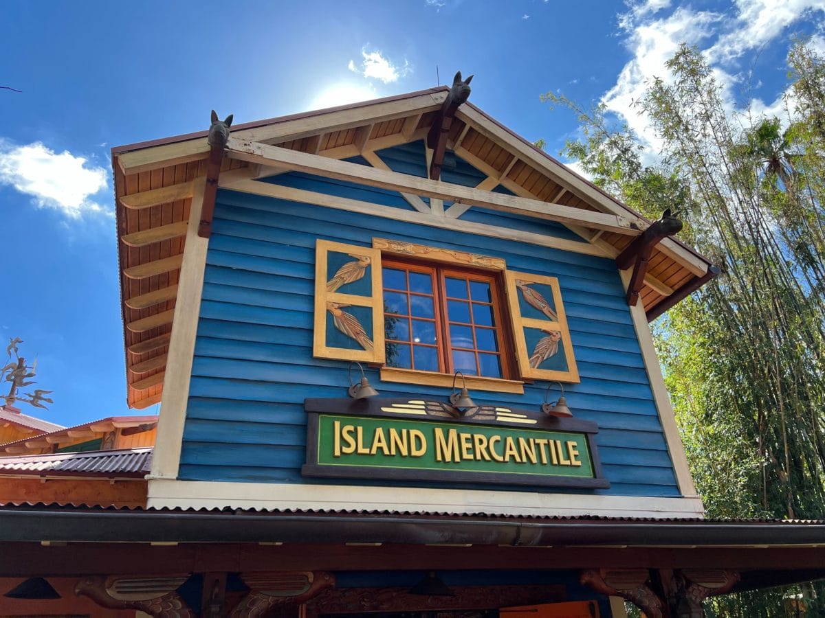 Blue wooden Island Mercantile shop with yellow-trimmed window at Disney World, vibrant under clear skies—no castle in view.