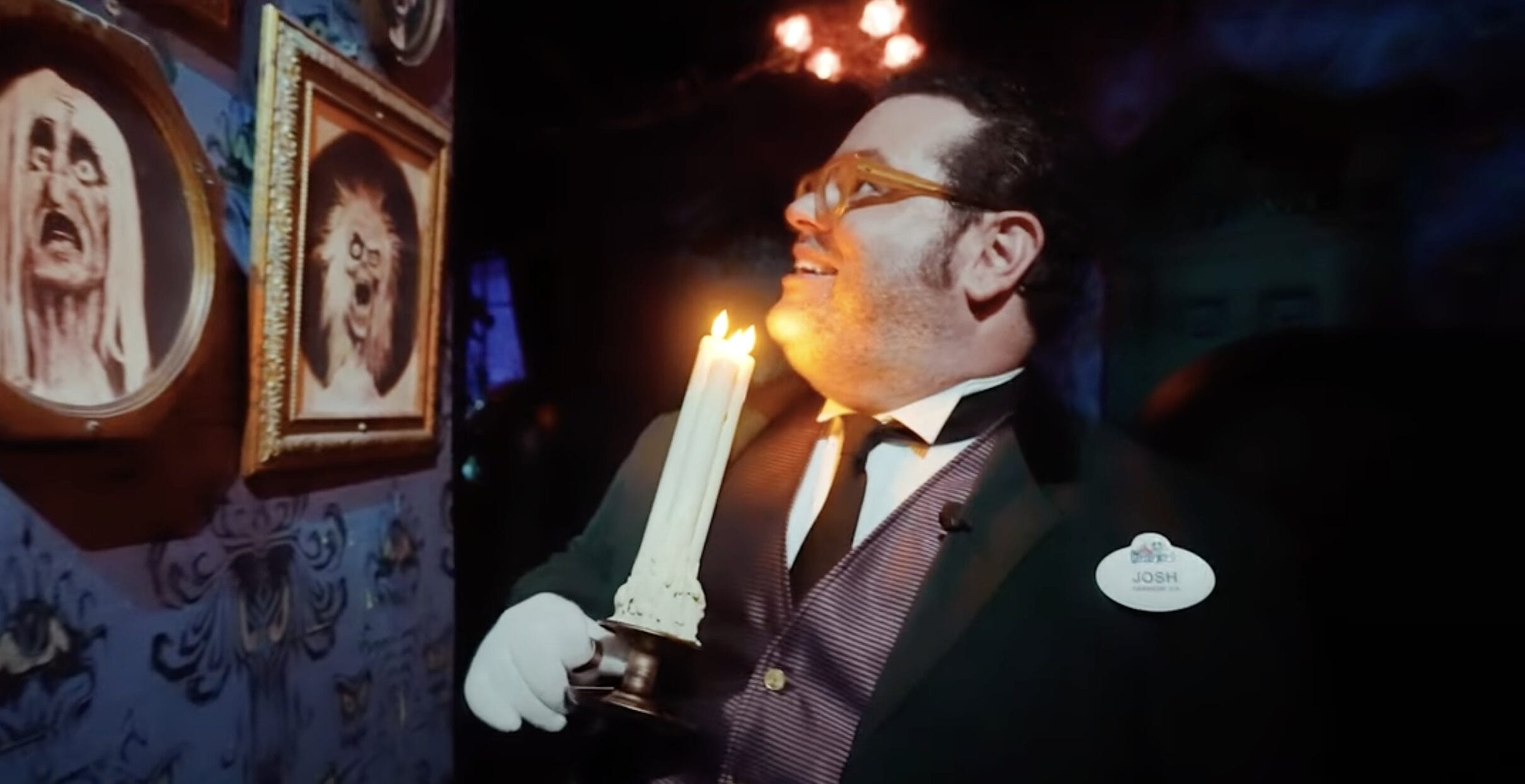 A man in a suit holding a candle gazes at eerie portraits on a dimly lit Haunted Mansion wall.