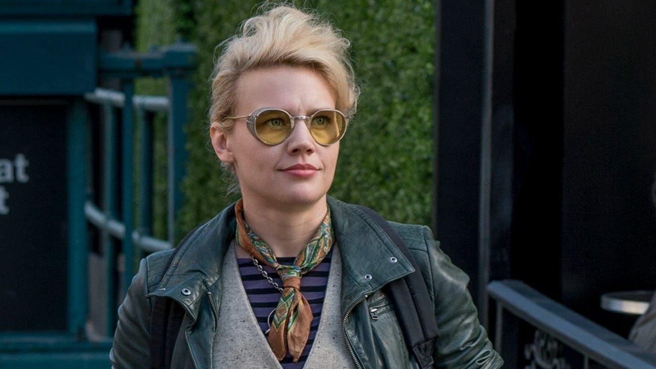 Person with short blond hair, round yellow glasses, and scarf, Kate McKinnon style, stands at Disney, no castle in sight.