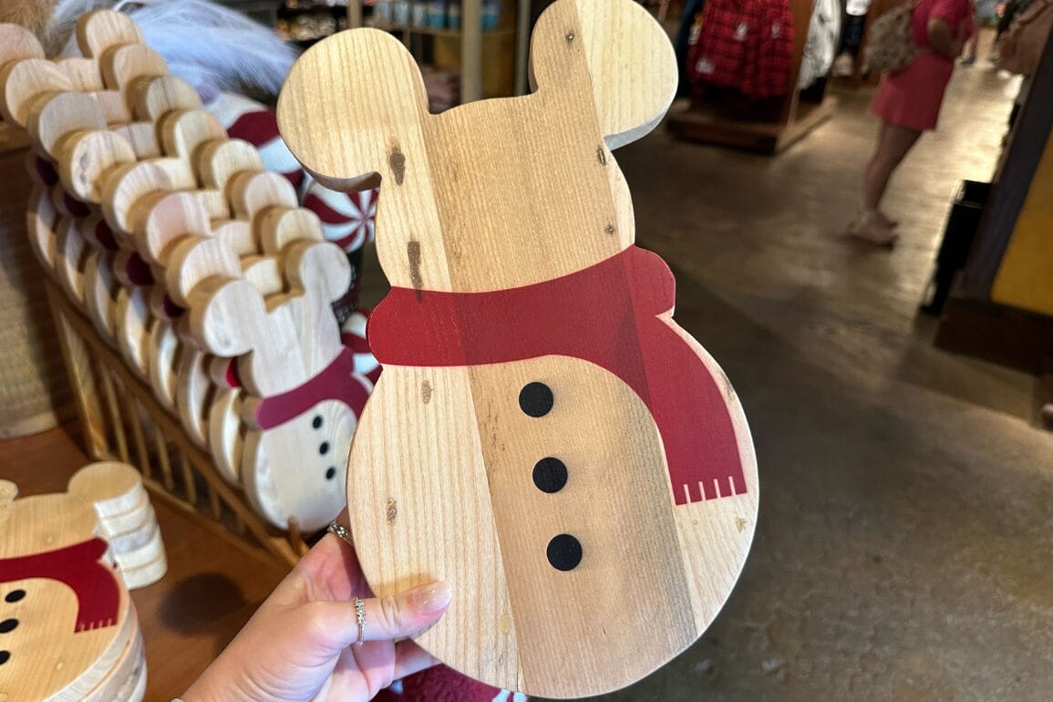 دستی که یک آدم‌برفی چوبی با گوش‌های میکی، شال‌گردن قرمز و دکمه‌های سیاه را نگه داشته است—یادآور سوغاتی‌های جشن در پارک‌های دیزنی.