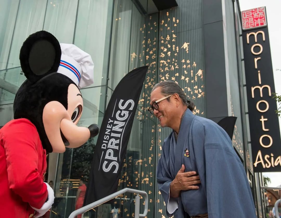 Mickey Mouse in a chef outfit bows to a smiling man outside Morimoto Asia at Disney Springs’ 10th Anniversary celebration. No castle here, so no comparison with Cinderella Castle at Walt Disney World or other Disneyland castles needed.