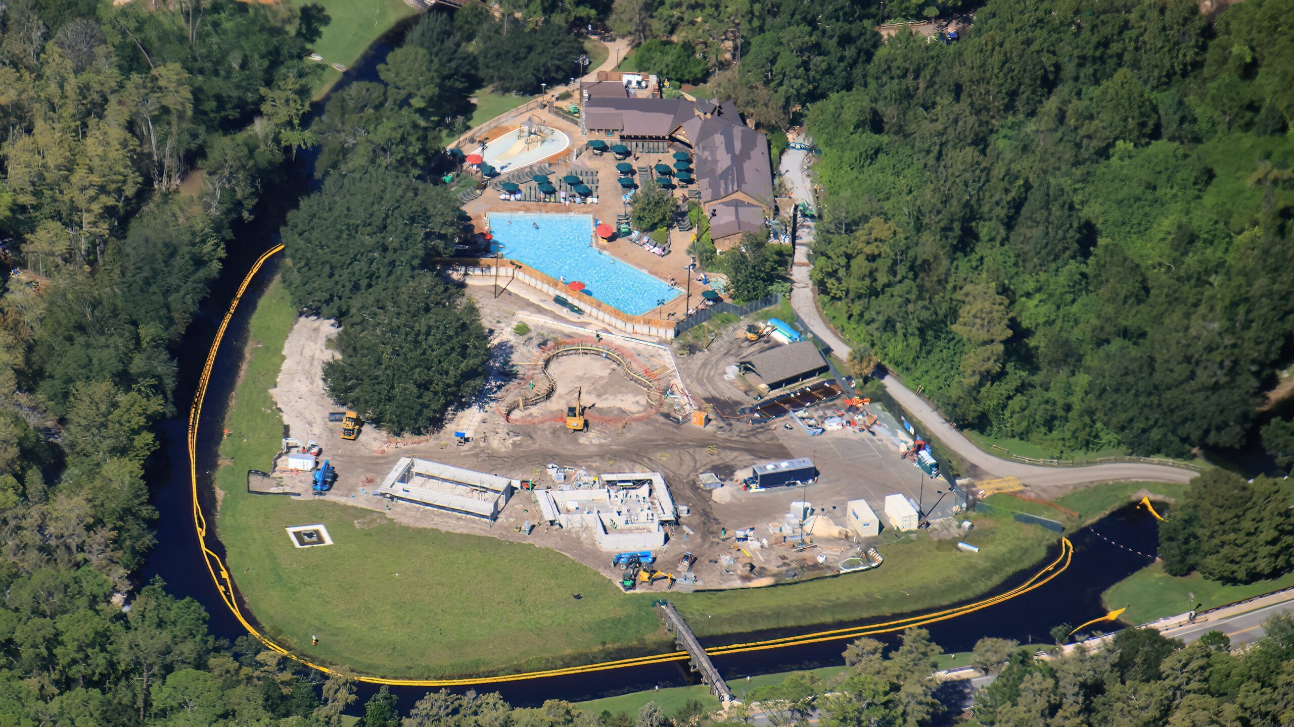 Aerial view of a construction site near a Disney resort with pools, lush trees, and a winding river, reminiscent of large Disney parks.