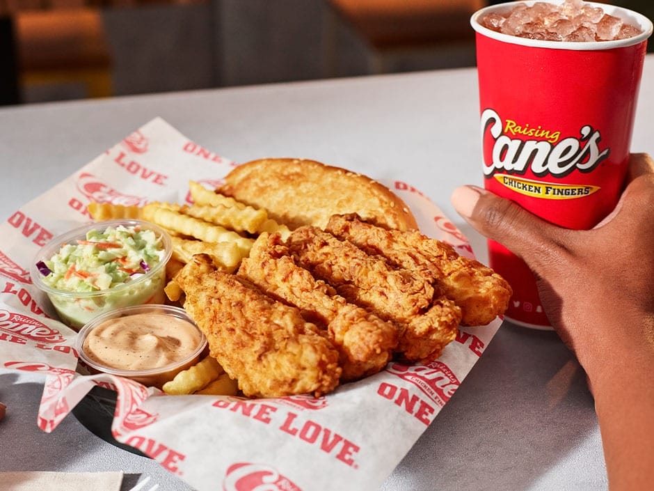 A plate of chicken fingers, crinkle fries, coleslaw, toast, and sauce beside a hand holding a Raising Cane’s cup at Disney park.
