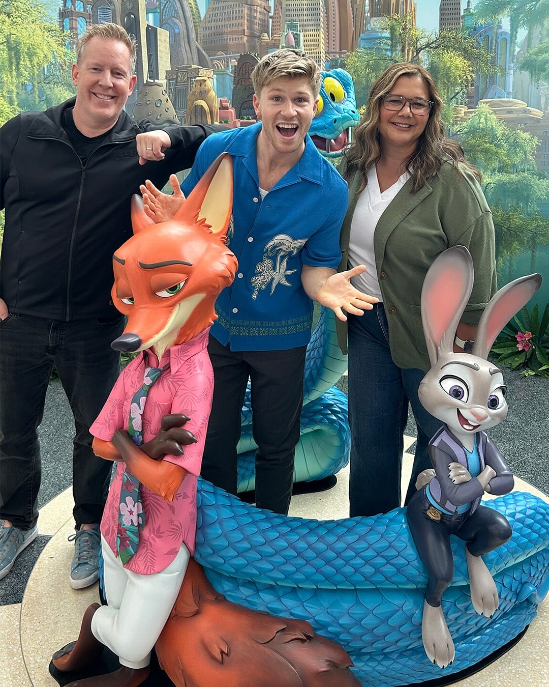 Three adults with Nick Wilde and Judy Hopps statues at a Disney park, a blue cartoon snake coiled behind them—no castle in view.