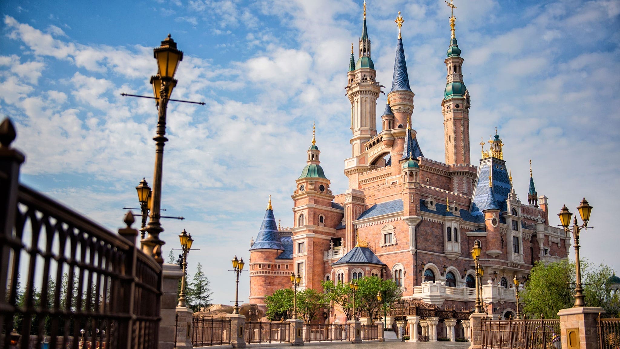 A castle with blue rooftops and golden spires, resembling Disneyland’s Sleeping Beauty Castle, stands beneath a partly cloudy sky.