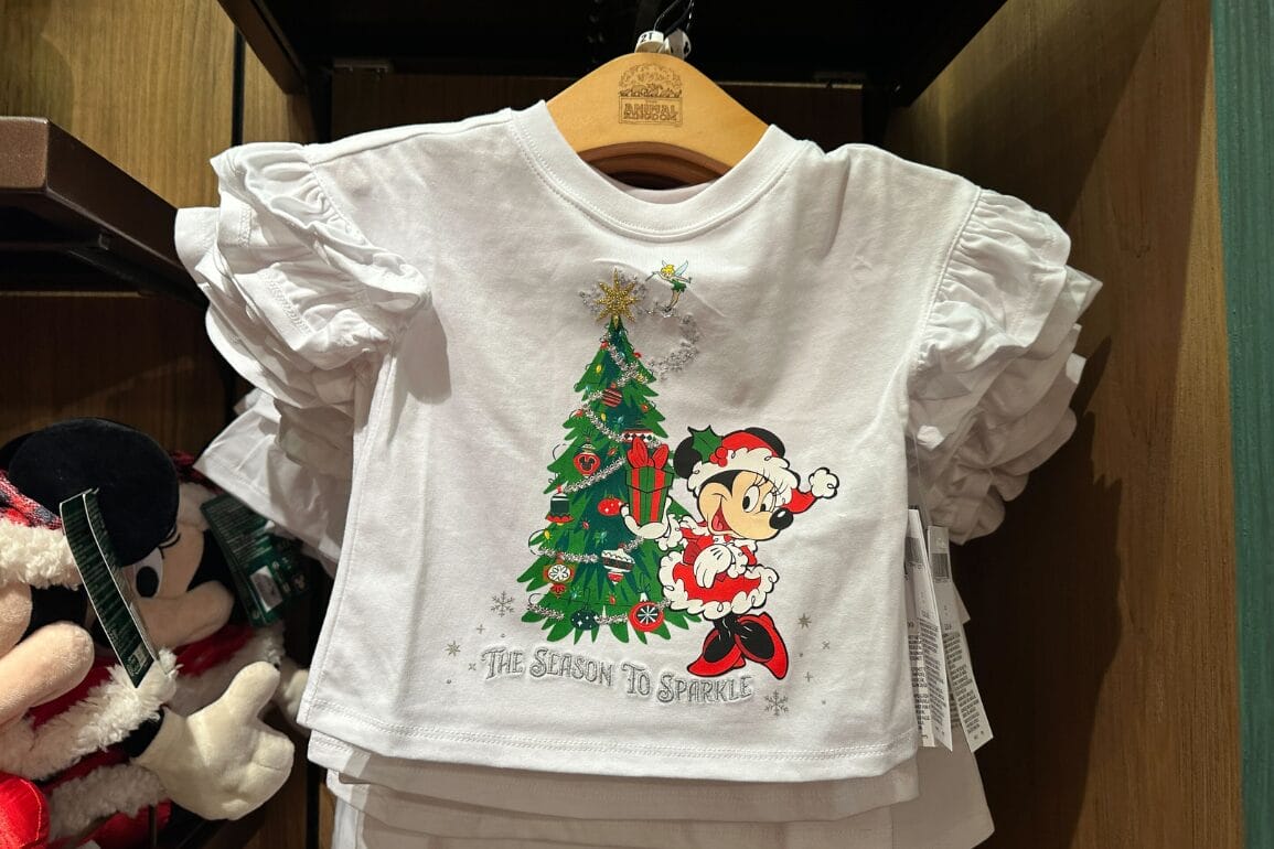 تی‌شرت سفید بچه‌گانه با طرح مینی ماوس در کنار یک درخت کریسمس، استایلی شاد که در پارک‌های دیزنی در فصل تعطیلات فروخته می‌شود.