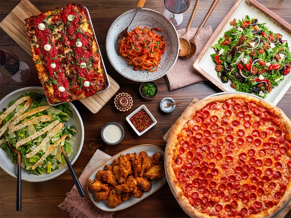A table topped with pepperoni pizza, lasagna, wings, salads, and pasta, reminiscent of Disney park dining but without castle views.