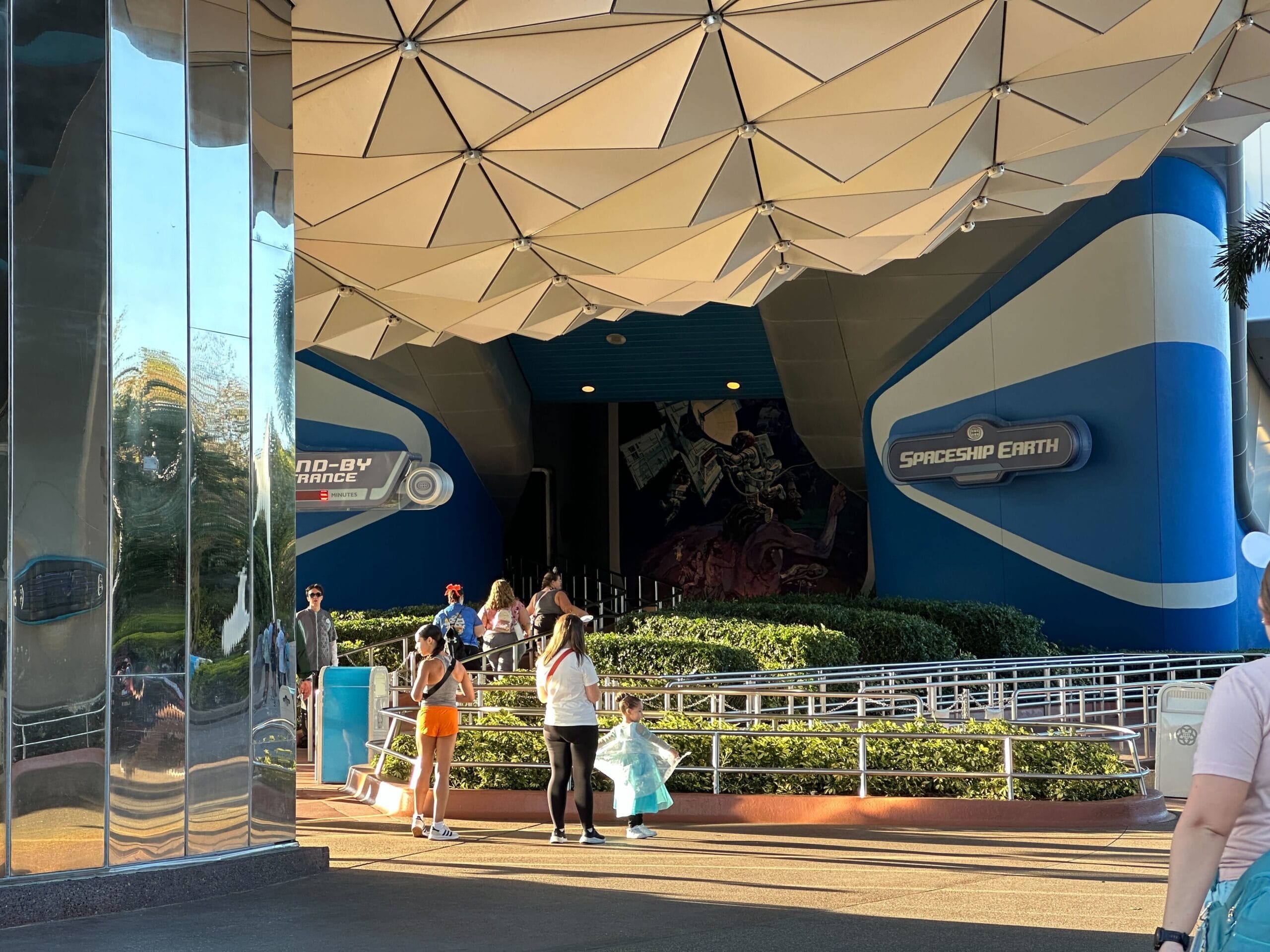Guests stroll by EPCOT's Spaceship Earth entrance, blue refurbishment signs visible, with its iconic silver geodesic sphere above.