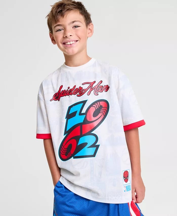 Smiling boy at a Disney park in a white Spider-Man 1962 Macy's shirt and blue shorts, hand in pocket; no Disney castle visible.