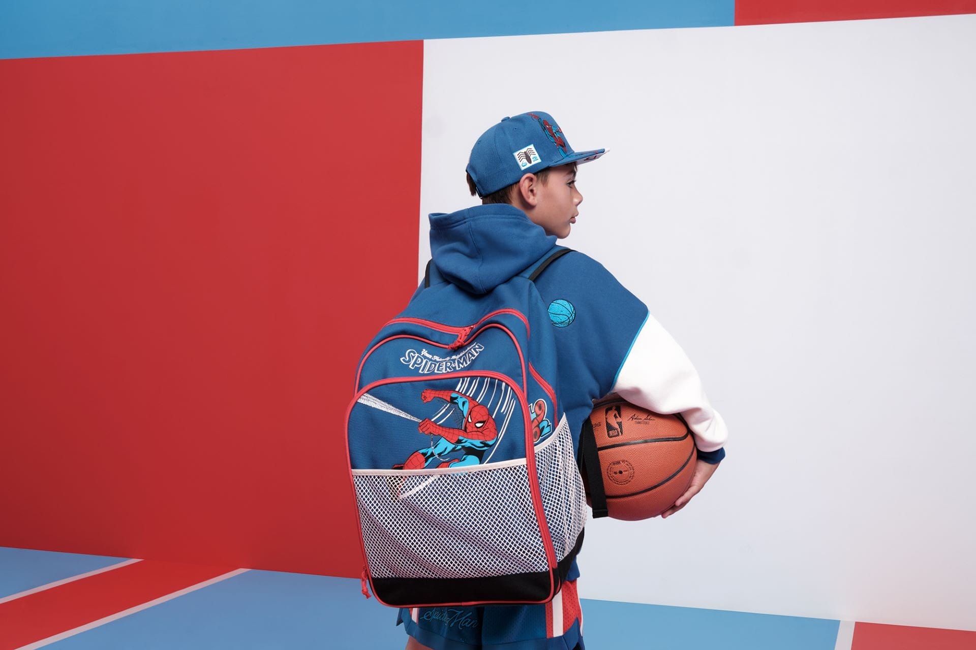 A boy in blue sportswear with an NBA basketball and Spider-Man backpack stands in a vibrant Disney-themed room.