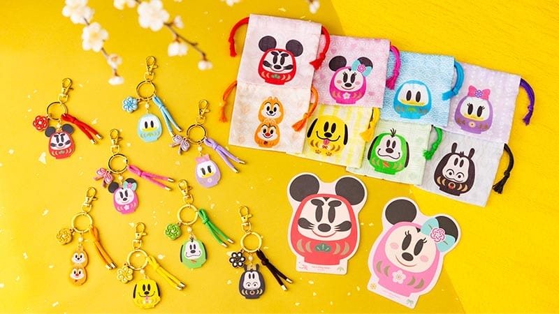 Disney Parks-inspired daruma charms, keychains, and pouches on a yellow background with floral accents, no castles pictured.