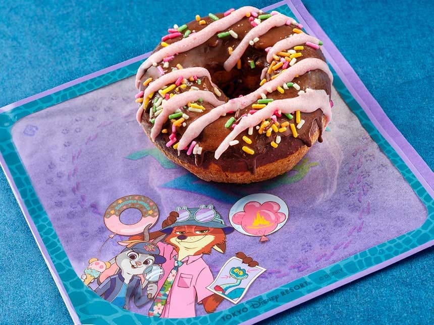 A chocolate donut with pink icing rests on a Tokyo Disneyland napkin with animated characters, no castle in view for comparison.