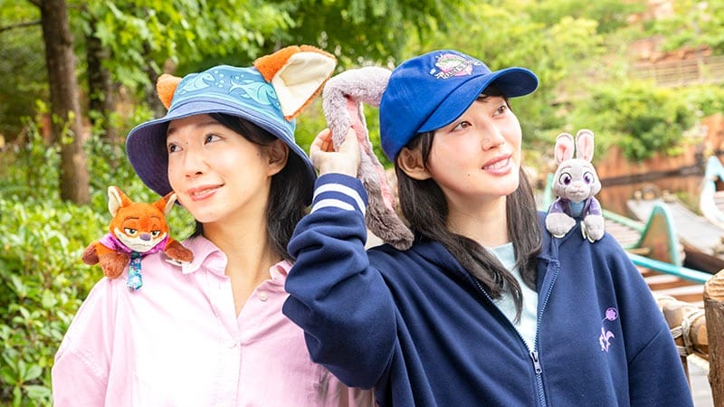 Two women in animal ear hats show Zootopia 2 merchandise, smiling at Tokyo Disneyland with greenery, no castle visible behind them.