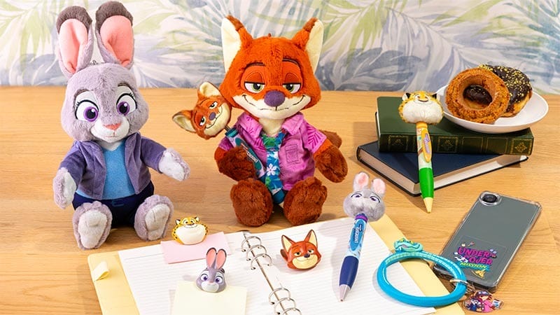 Zootopia 2 plush toys, stationery, and themed pastries cover a desk beside books and a notepad, like Disney park merch displays.
